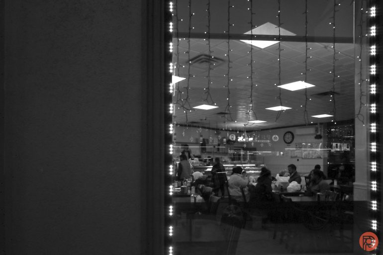 People dining inside a restaurant, viewed through a window decorated with string lights at night.