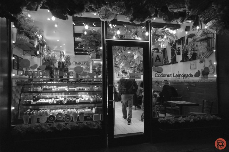 A person stands inside a cozy bakery at night, with pastries displayed in the window and hanging decorations.