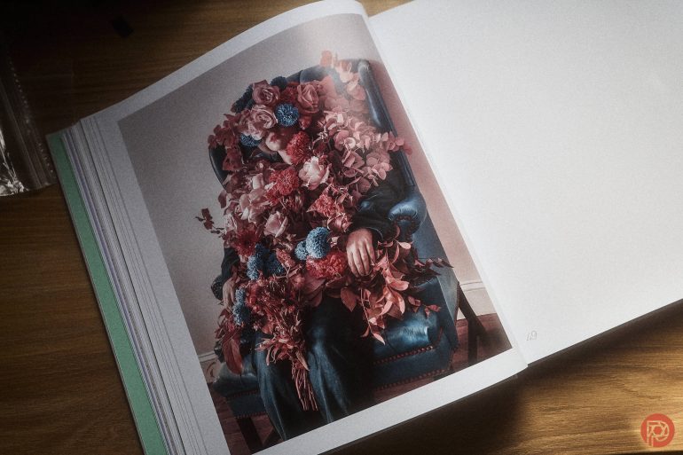 A book open to a photo of a person covered in colorful flowers sitting in a blue chair.