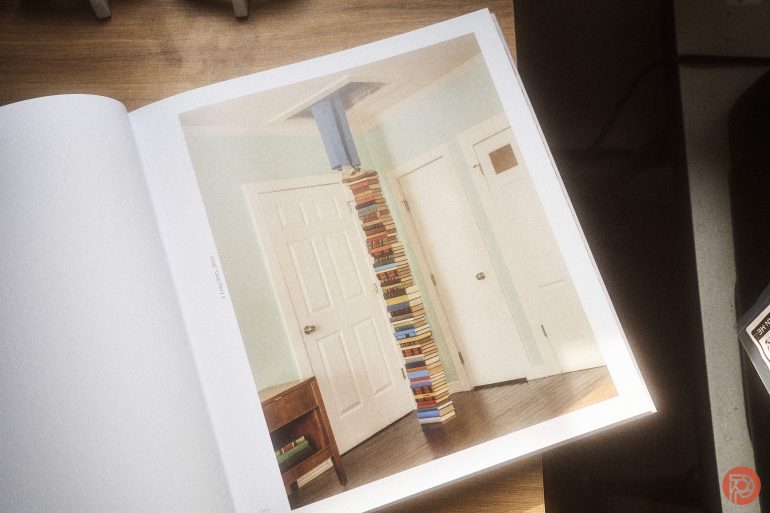 An open book shows a photo of a tall, vertical stack of books reaching the ceiling in a room with white doors.