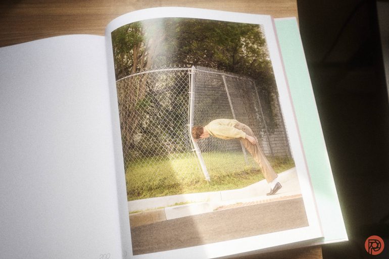 A person in beige clothes leans forward at a sharp angle by a chain-link fence near greenery.