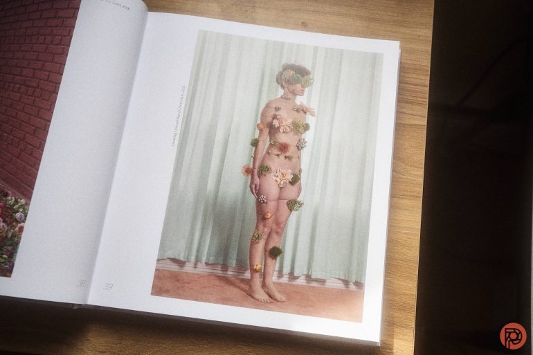 An open book shows a photo of a nude person standing, covered with flowers, against a pale curtain background.