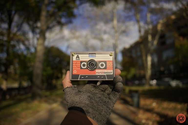 A gloved hand holds a cassette-shaped camera outdoors on a sunny, tree-lined path.