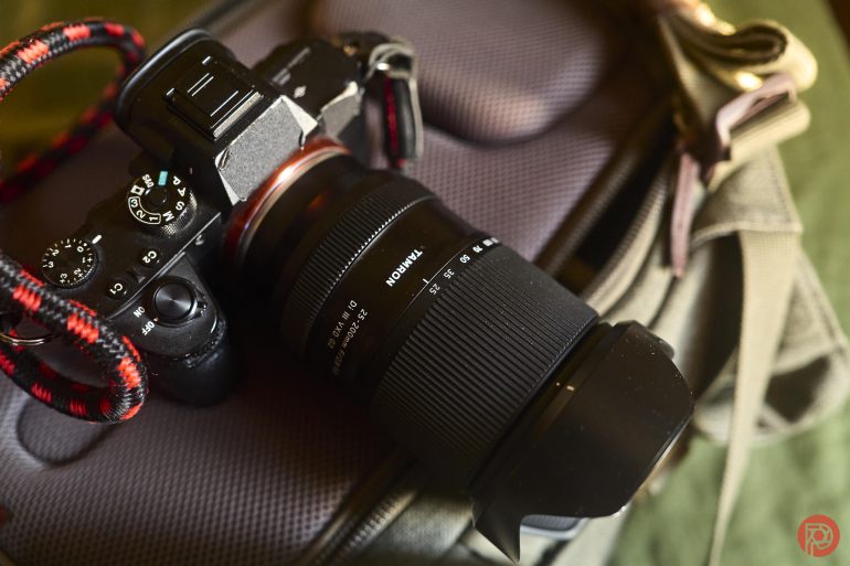 A camera with a zoom lens rests on a gray camera bag with a red-black strap attached.