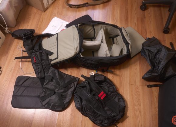 A camera backpack with open compartments and accessories spread out on a wooden floor.