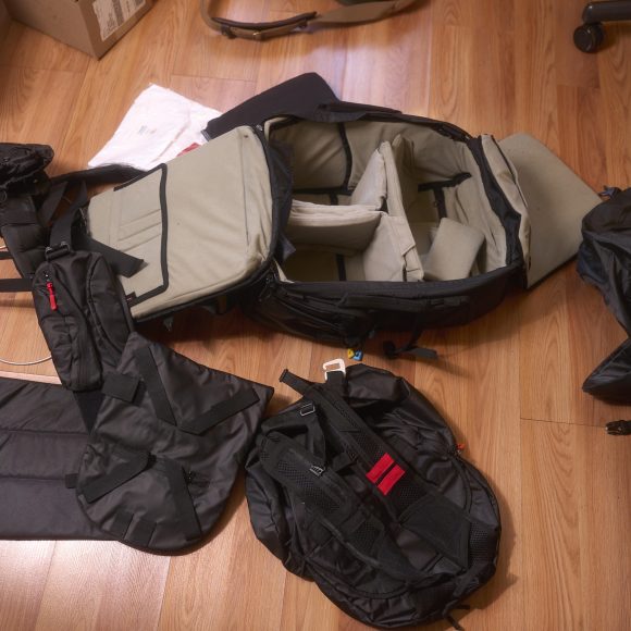 A camera backpack with open compartments and accessories spread out on a wooden floor.