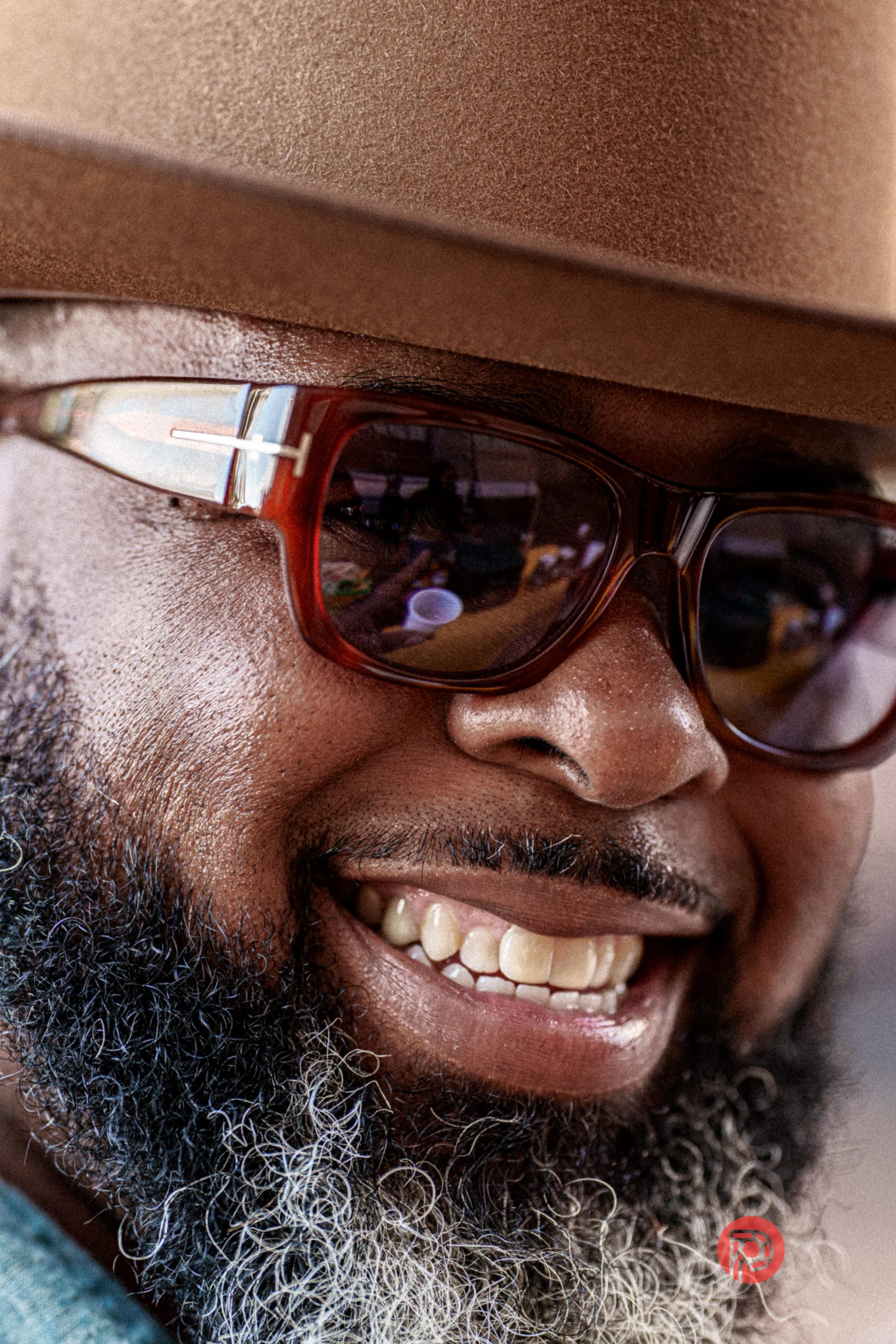 A smiling man with a beard, wearing a brown hat and red sunglasses, looks directly at the camera.