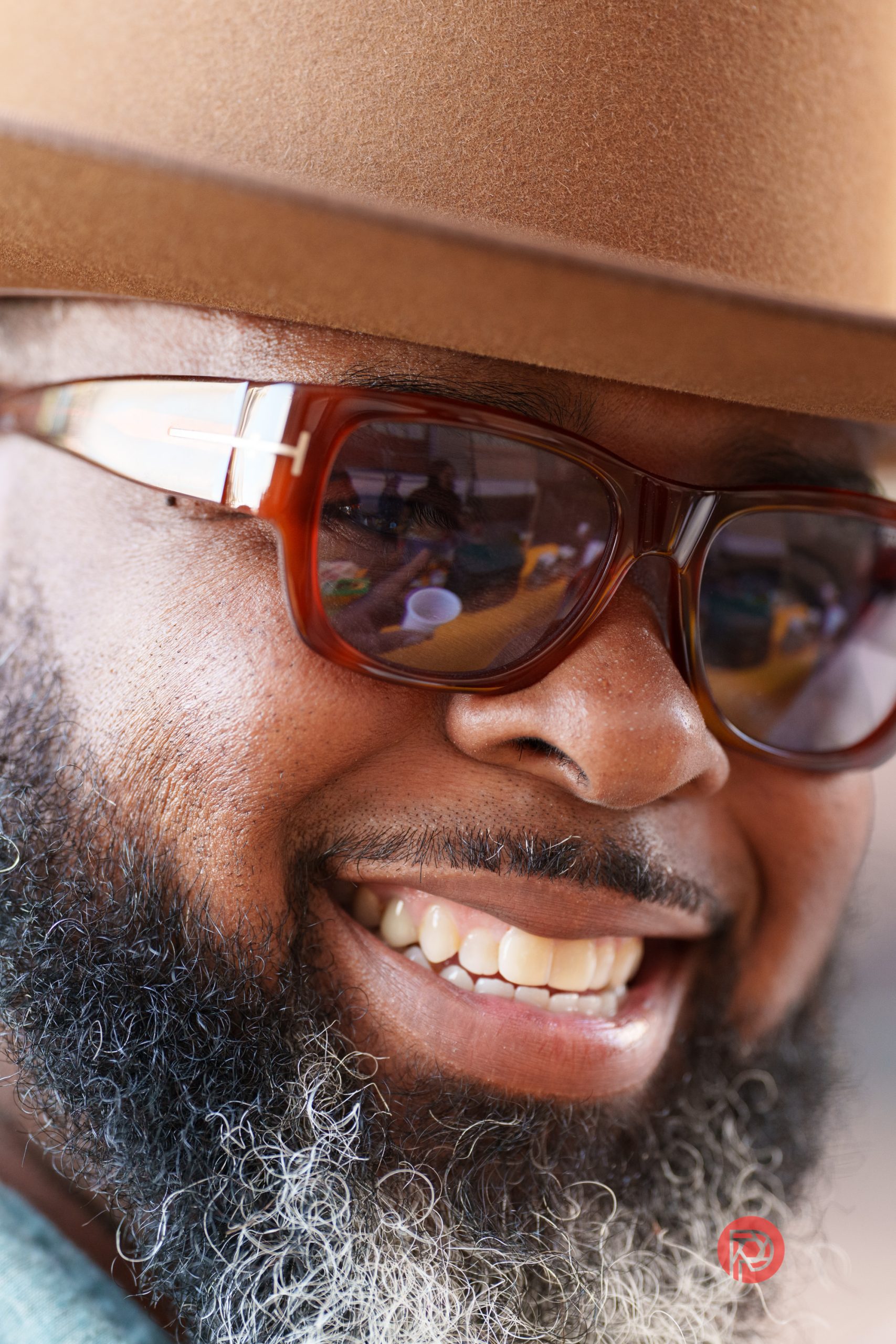 Smiling man with a beard in sunglasses and a brown hat, showing his teeth.