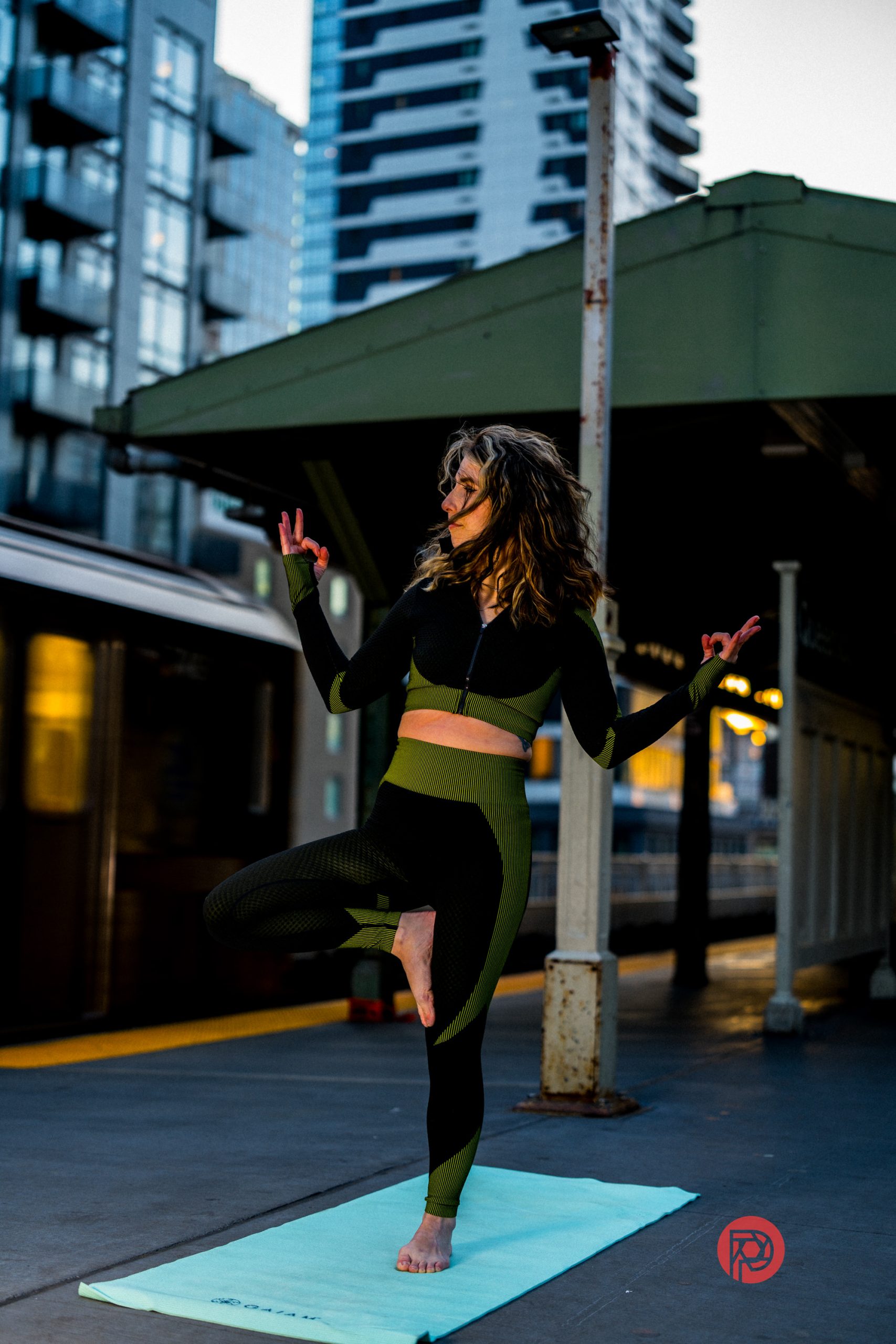A woman in activewear does a tree yoga pose on a mat at an urban train station platform.