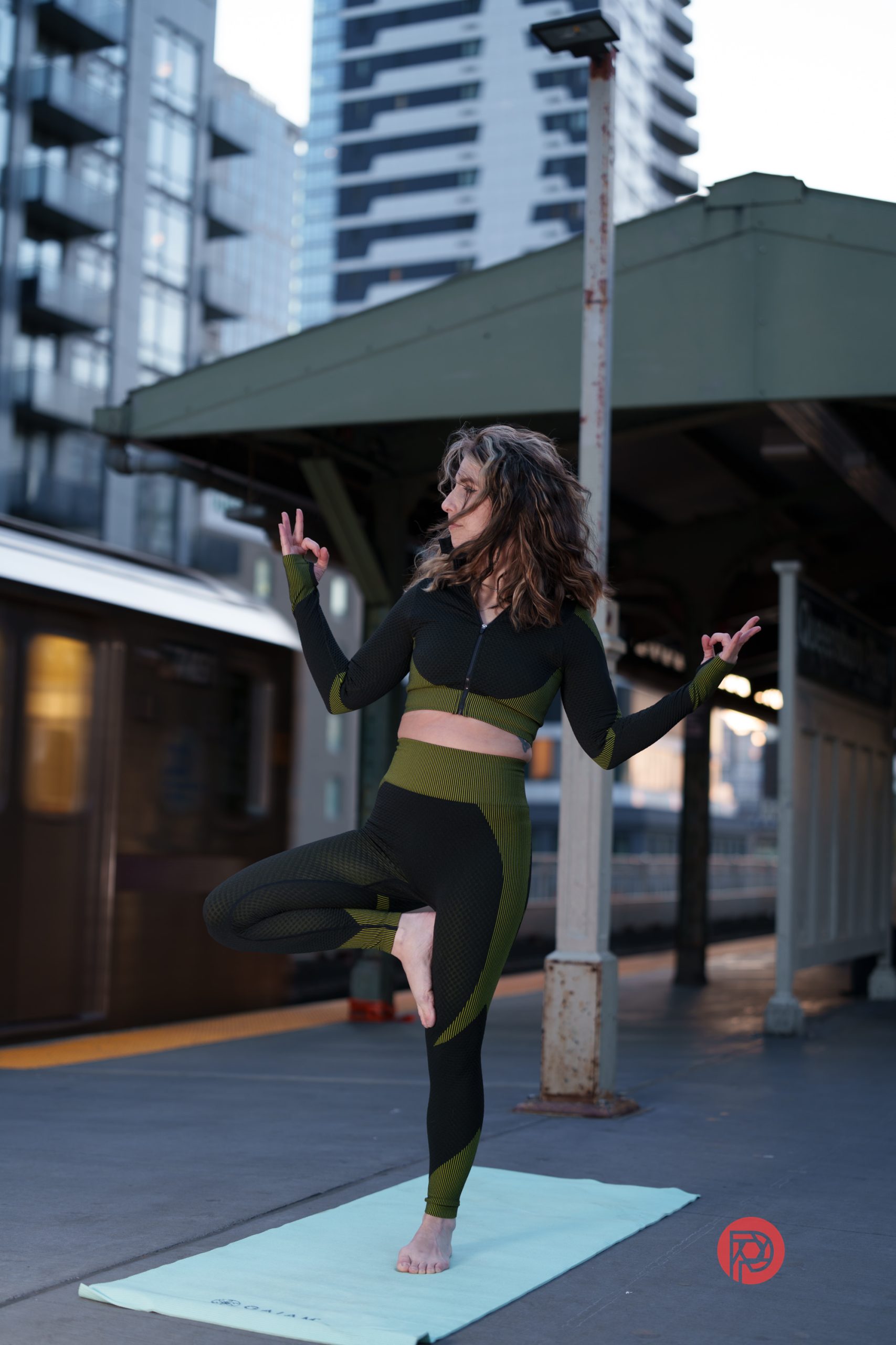 Woman in athletic wear does a yoga pose on a mat at an urban train station with tall buildings in background.