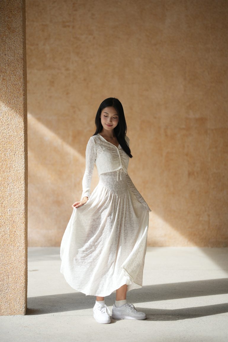 Woman in a light, flowing dress standing in sunlight against a textured beige wall, looking down and smiling.