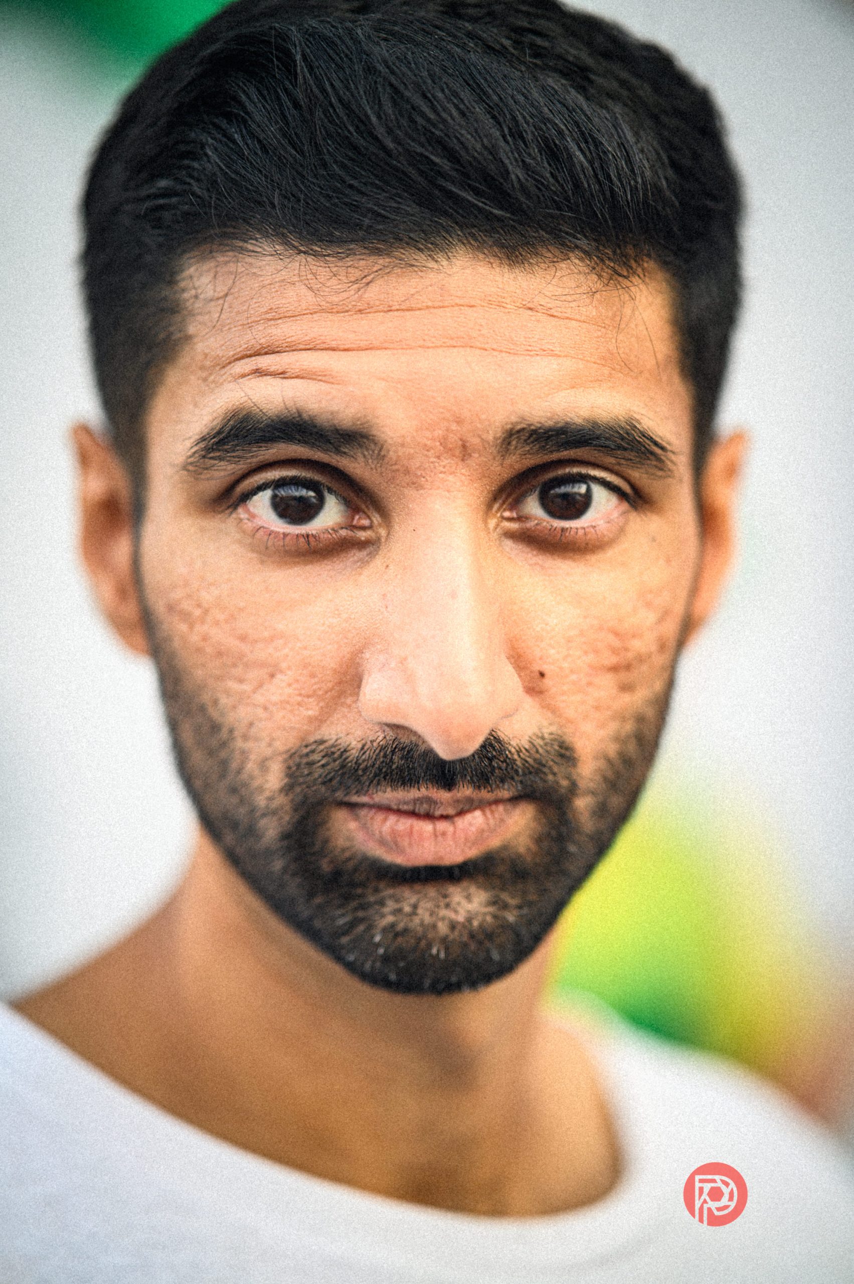 A man with short dark hair and facial stubble looks directly at the camera, wearing a white shirt.