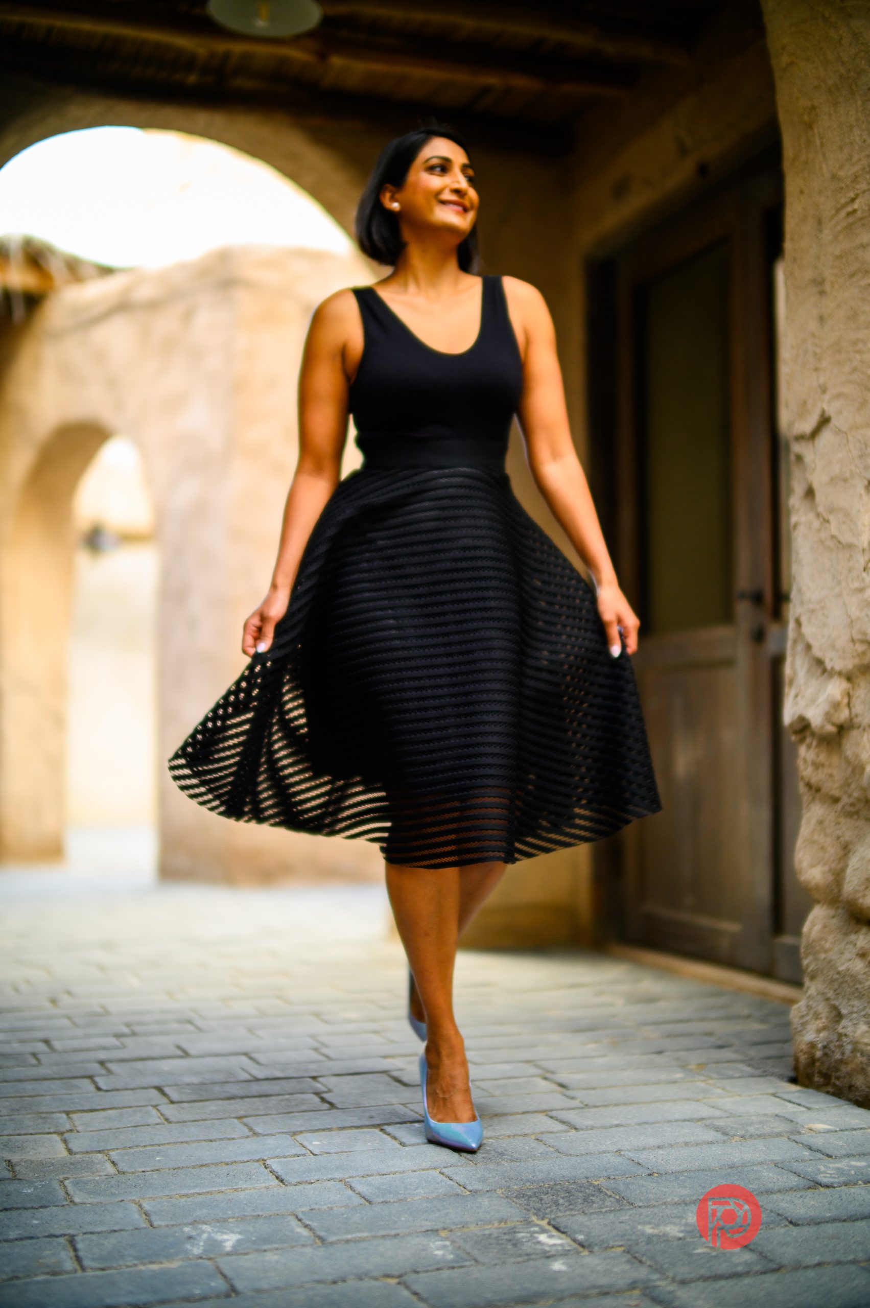 Woman in a sleeveless black dress and heels walking and smiling in a rustic, sunlit outdoor corridor.
