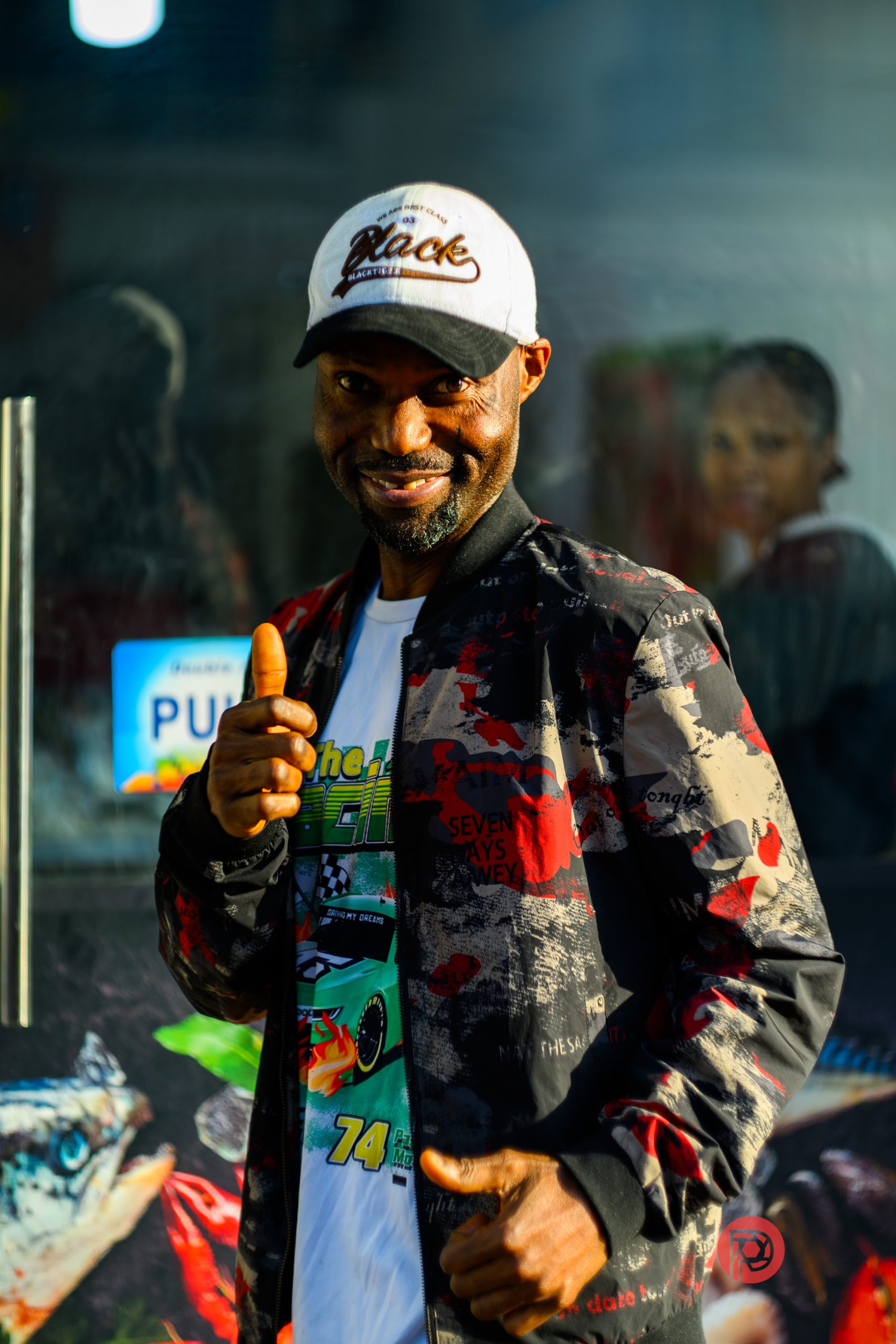 A smiling man in a patterned jacket and white cap gives a thumbs up, standing outdoors on a sunny day.
