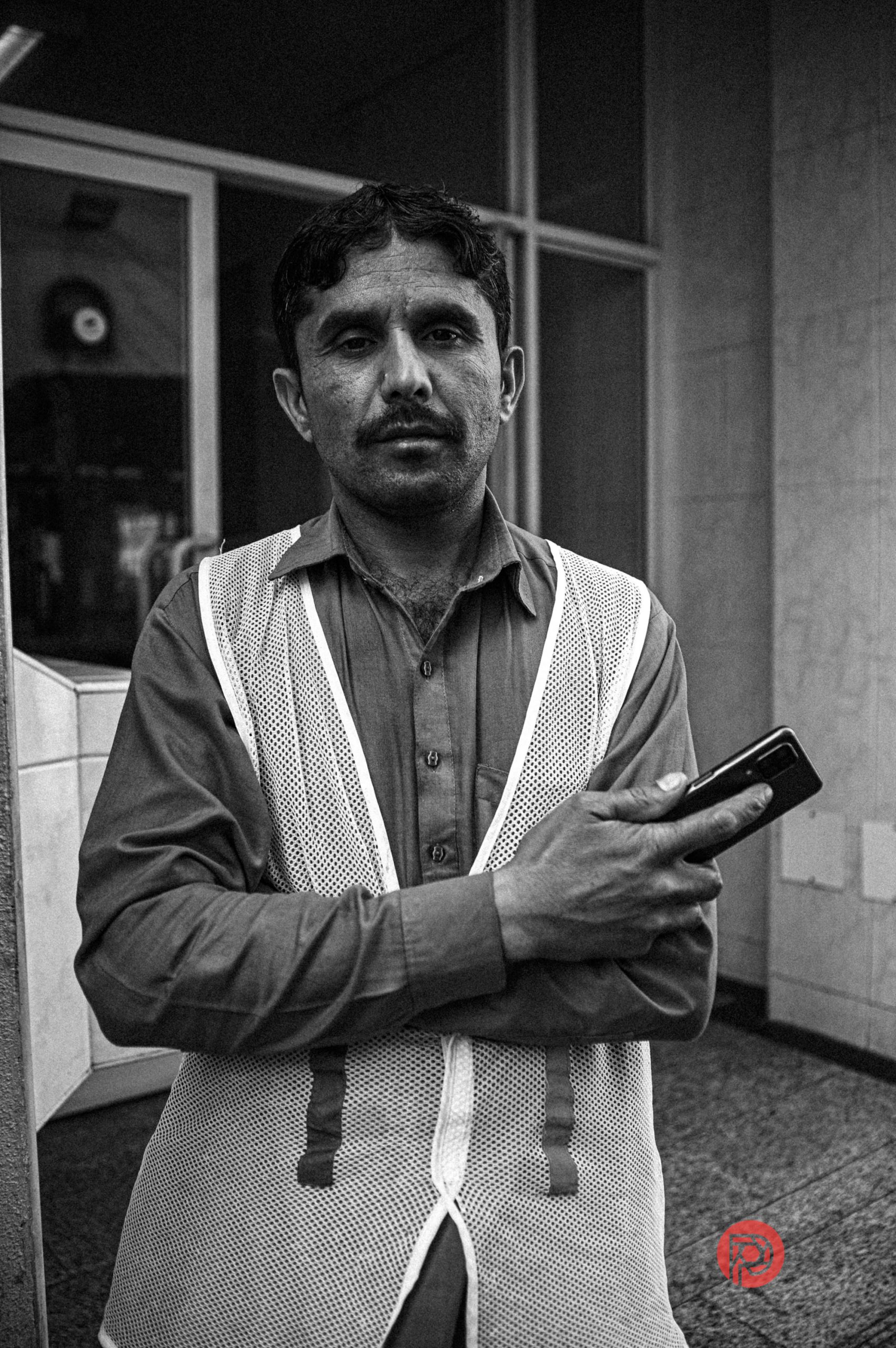 A man in a mesh vest stands with folded arms holding a phone, looking at the camera in a city setting.