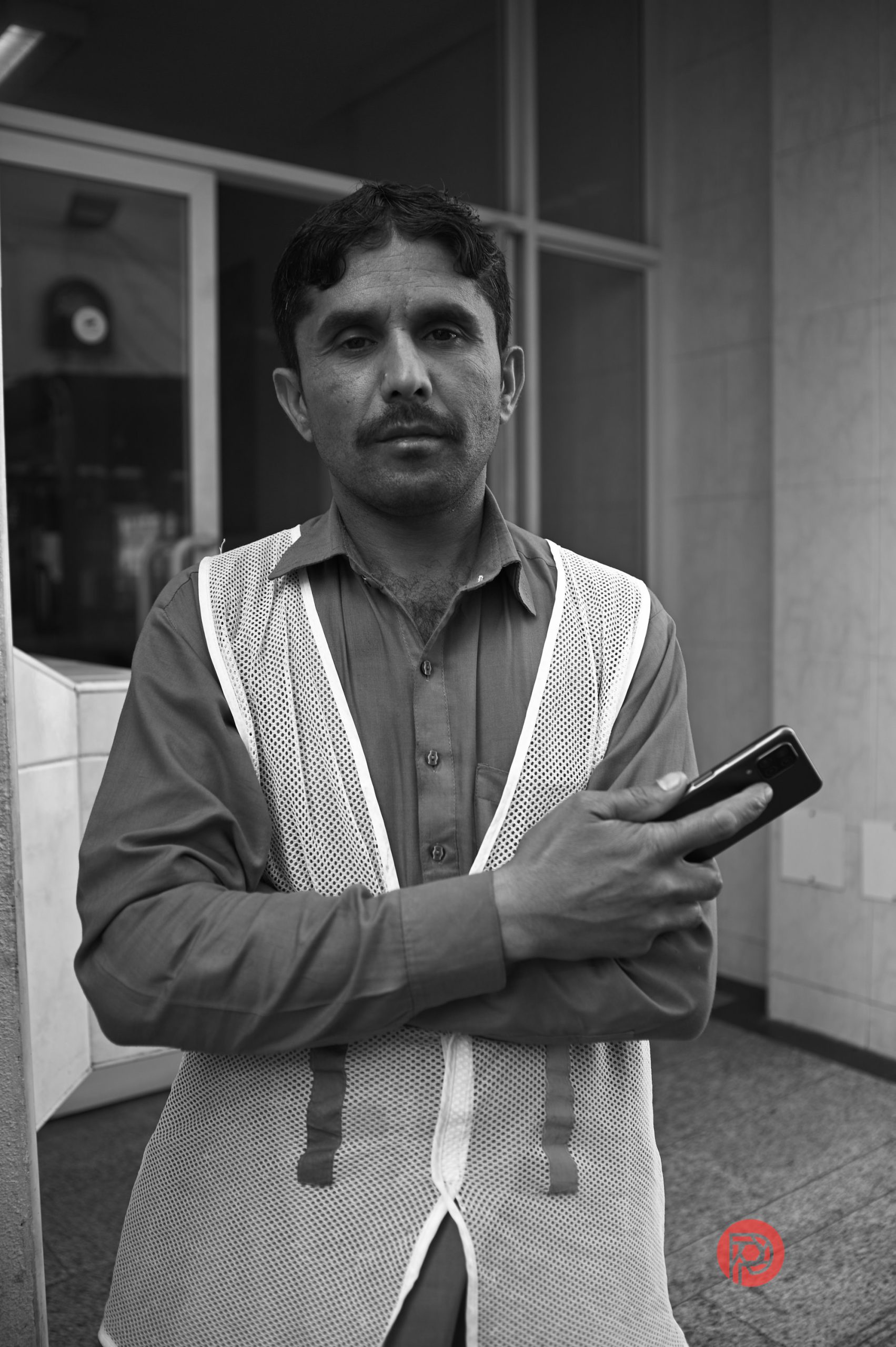 A man in a mesh safety vest stands with folded arms, holding a phone, outside a building.