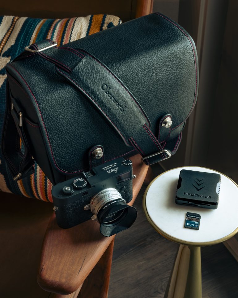 A black camera bag, a Leica camera, a memory card, and a card reader on a chair and small table.