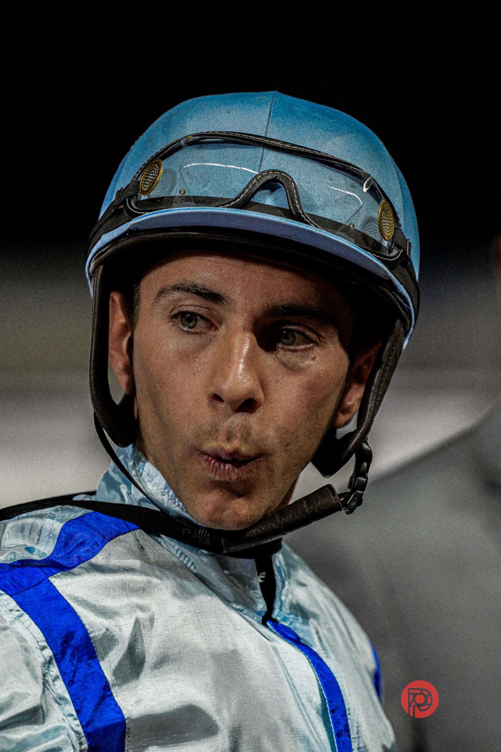 A jockey in blue and white gear with a helmet and goggles looks to the side with a surprised expression.