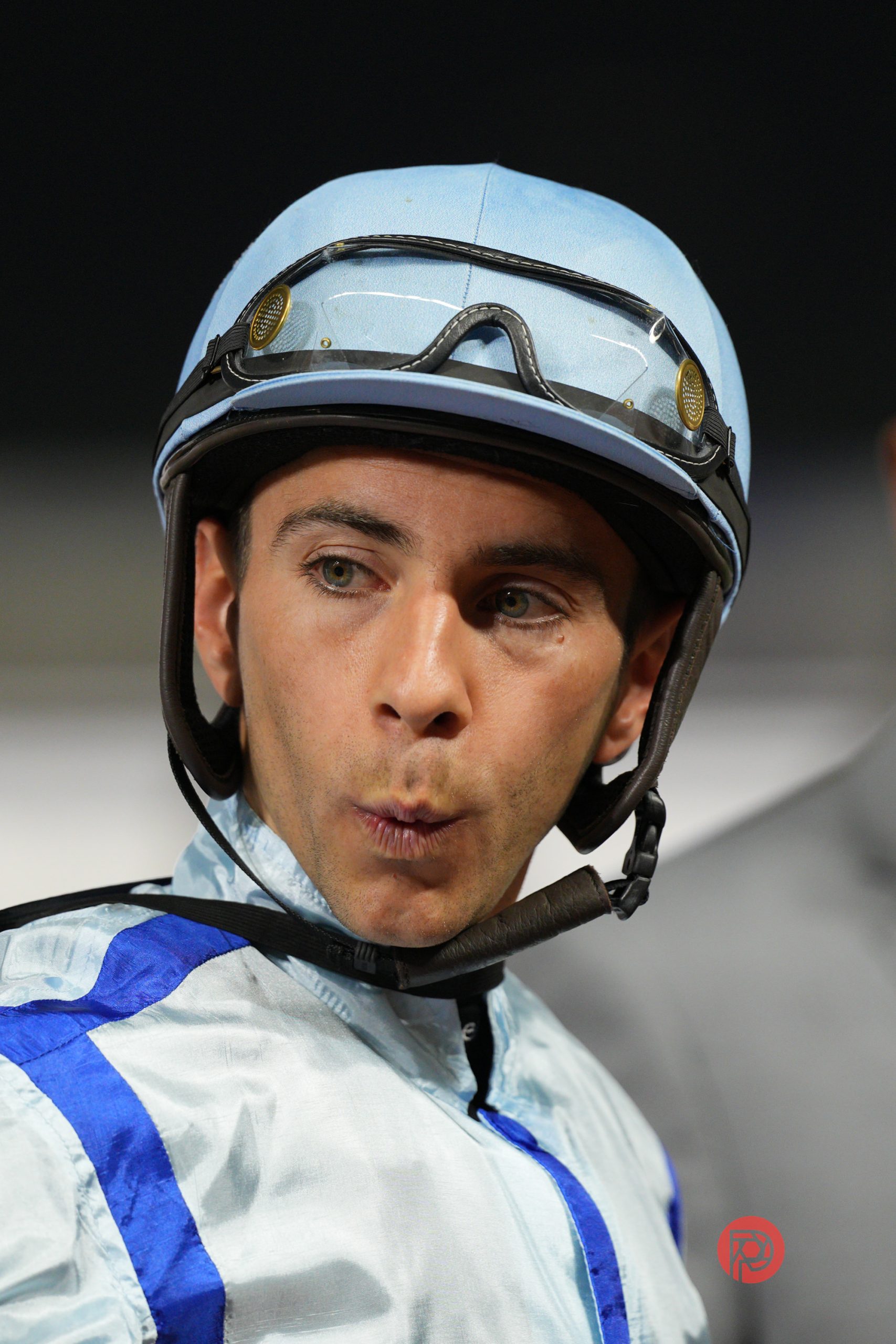 A jockey in a light blue helmet and silks looks surprised, with lips pursed and eyes wide.
