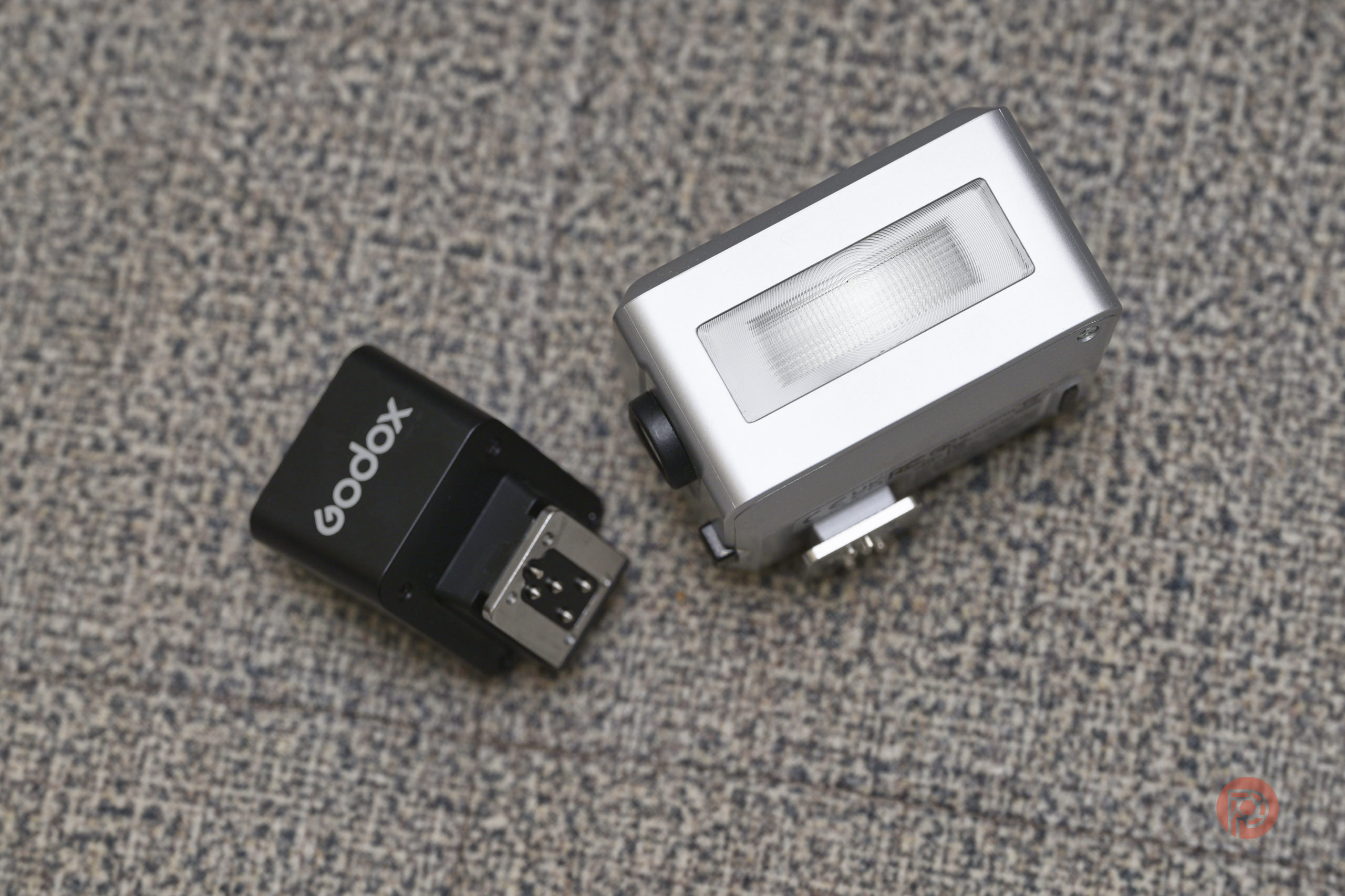 A Godox camera flash trigger and a small silver camera flash unit on a textured gray surface.