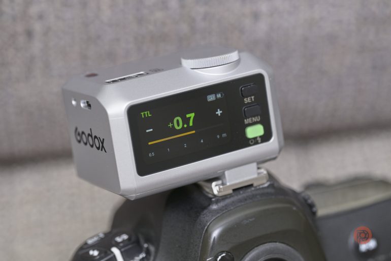 Close-up of a Godox camera flash trigger mounted on a camera, displaying flash settings on its screen.