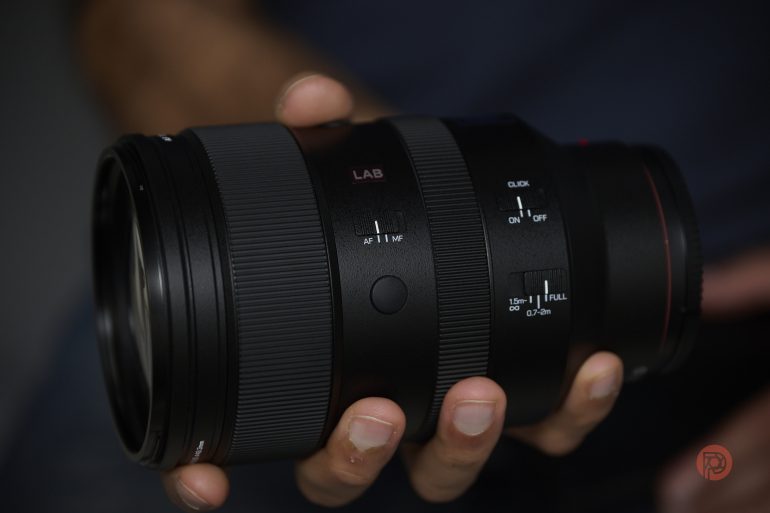 A hand holding a black camera lens with visible switches and textured focus ring.