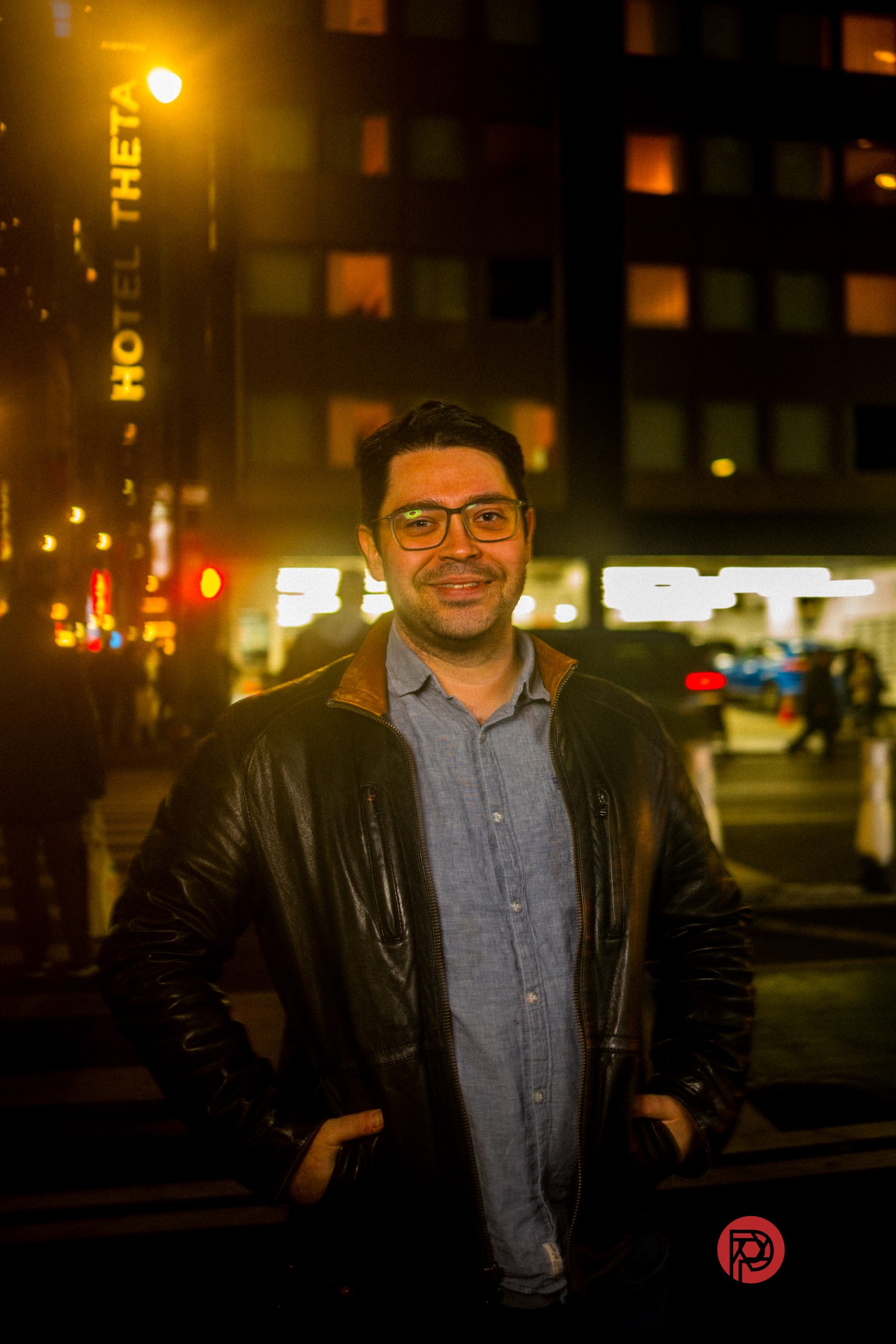 A man in glasses and a leather jacket stands smiling on a city street at night with bright lights behind him.