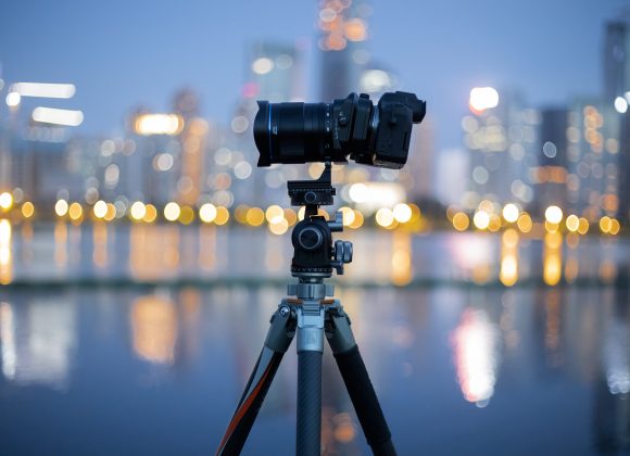 A camera on a tripod is set up by the water with a city skyline blurred in the background at dusk.