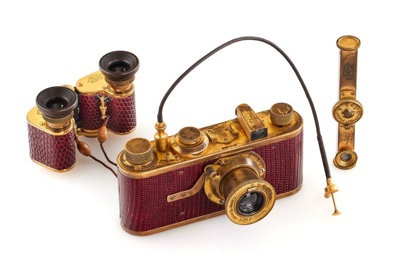 Vintage gold camera, matching binoculars, and rangefinder with red textured covering on a white background.