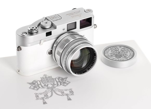 A silver Leica camera with a matching lens cap sits on a white surface with a decorative emblem.