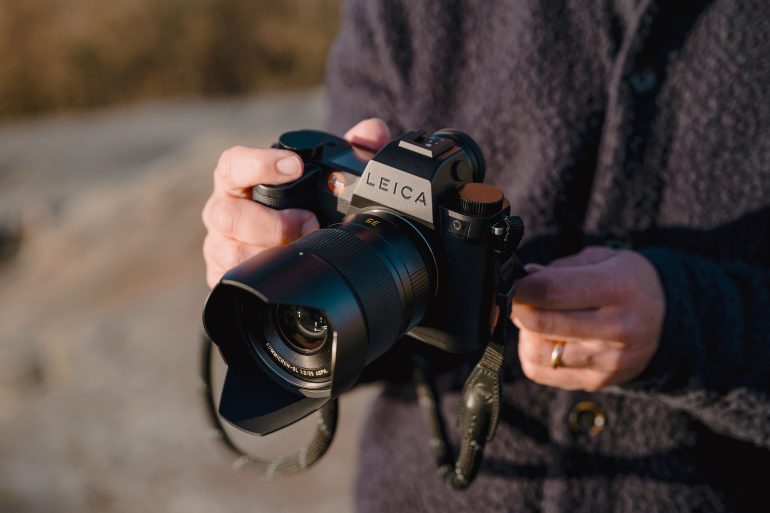 Person holding a Leica camera with a large lens outdoors, wearing a dark textured jacket.