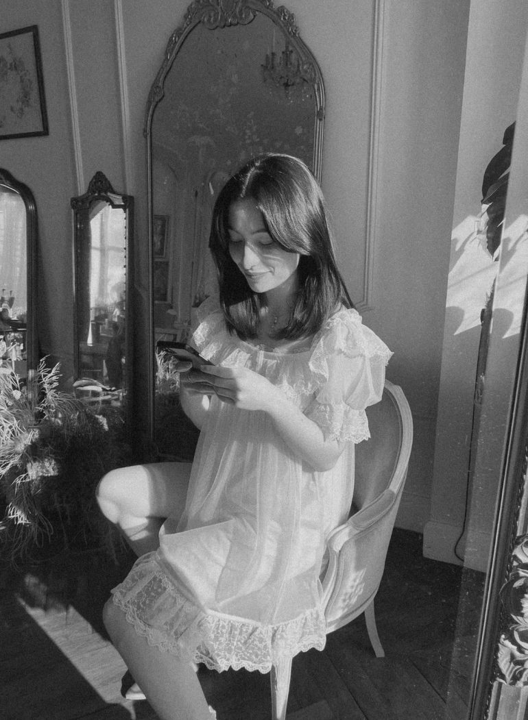 Woman in a white dress sits on a chair, looking at her phone, with mirrors and sunlight in the room.