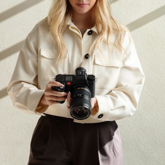 Woman in a cream jacket holding a professional camera, standing against a light-colored wall.