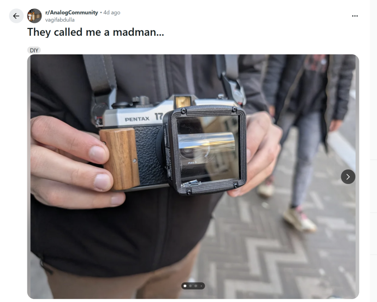 A person holds a Pentax camera modified with a large wooden grip and an attached viewfinder mirror.