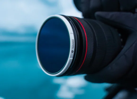 A gloved hand holds a camera with a lens filter, with a blurred blue background.