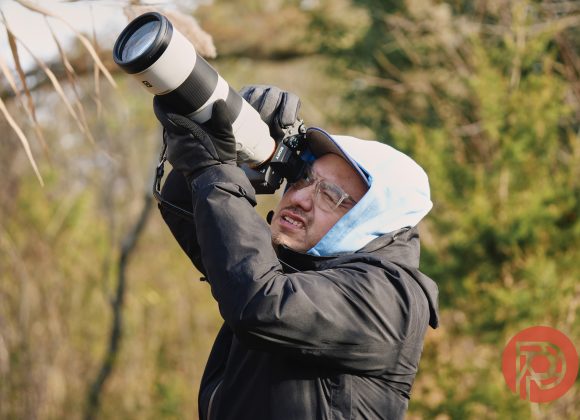 A person in a hooded jacket uses a camera with a large lens outdoors, aiming at the sky.