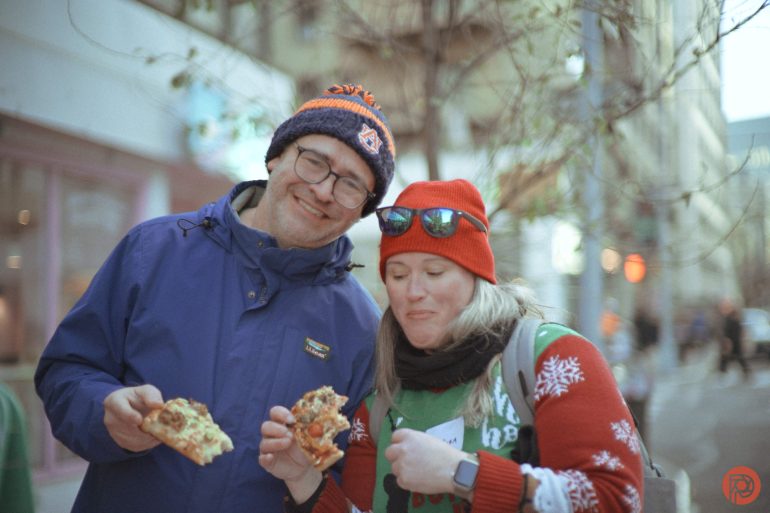 Two people in winter clothes smiling and eating pizza outside on a city street.