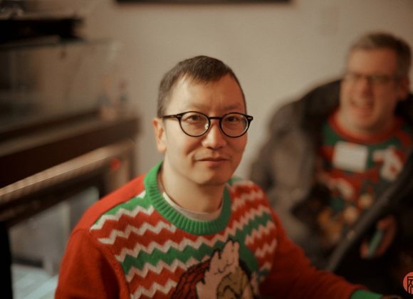 A person wearing glasses and a festive holiday sweater sits indoors, another person is blurred in the background.