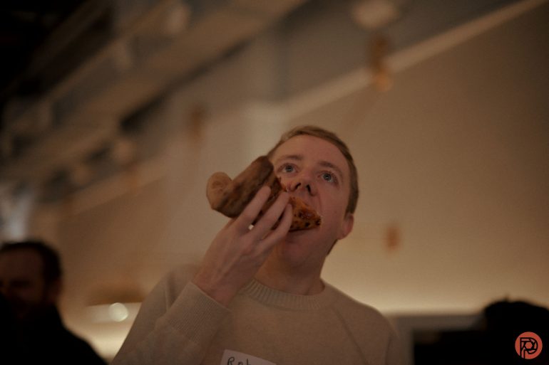 A man in a beige sweater eats a slice of pizza indoors under warm lighting.