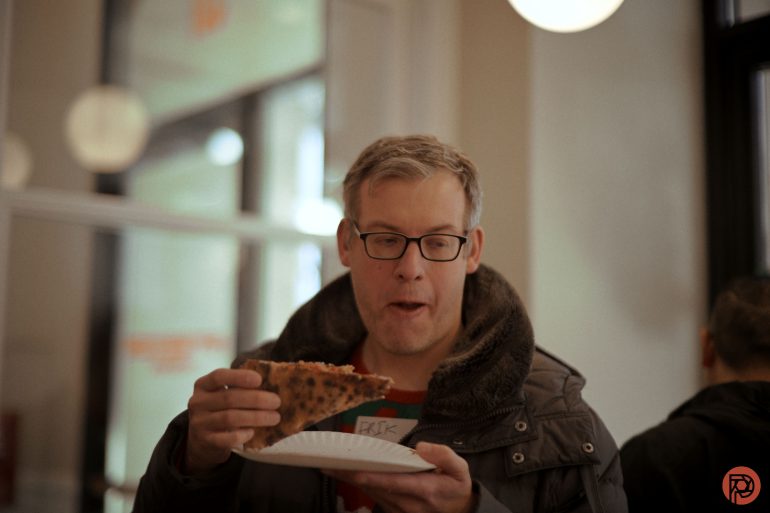 A man wearing glasses and a jacket holds a large slice of pizza and looks at it with interest.