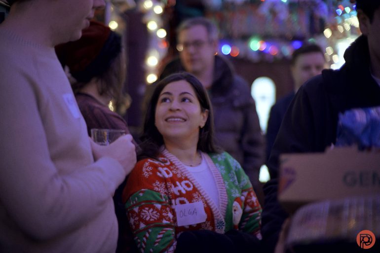 A woman in a festive sweater, smiling at a man, stands among people at a holiday party with string lights.