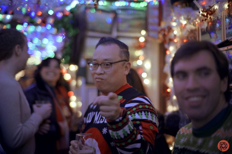A man in a festive sweater points at the camera at a holiday party with colorful lights and people around.