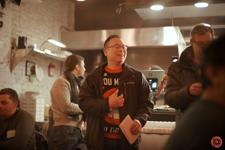 A man smiles while holding a microphone and paper in a cozy, warmly lit indoor setting with others around him.