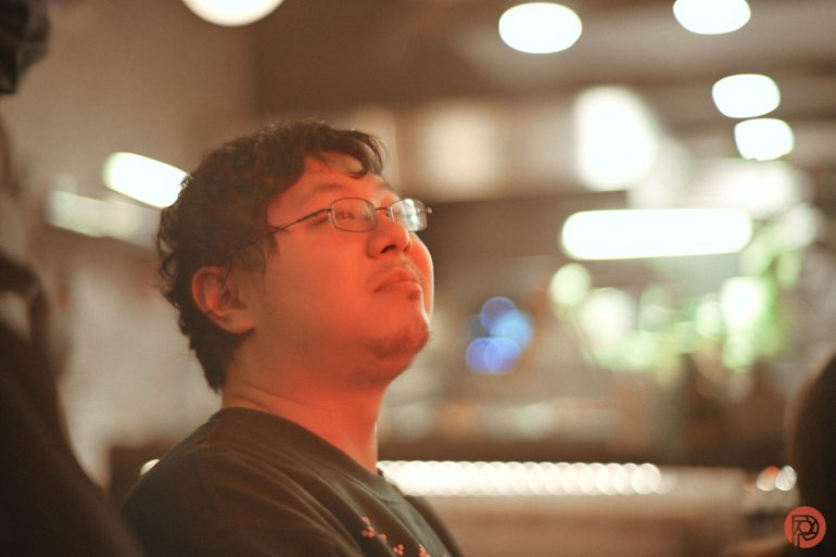 A man with glasses looks content under warm lighting in a softly blurred indoor setting.
