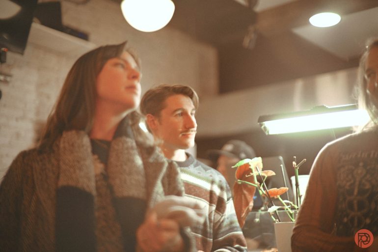Three people stand indoors near a lamp and orange flowers; the scene has a warm, soft focus.