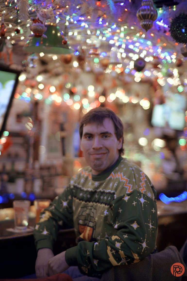 A man in a festive sweater sits at a bar with colorful holiday lights in the background.