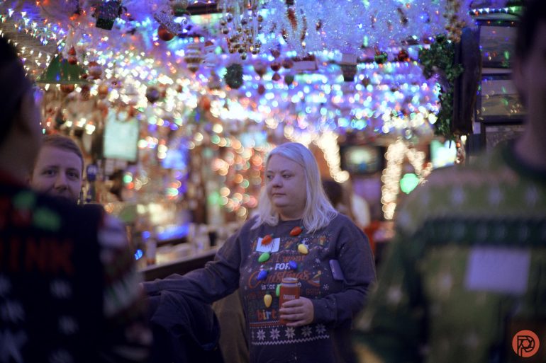 A woman in a festive sweater holds a drink in a bar decorated with colorful holiday lights.