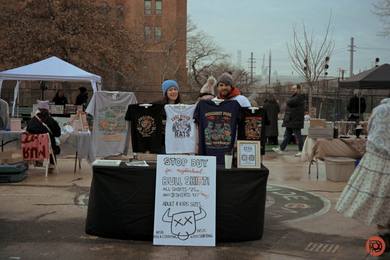Two people selling graphic t-shirts at an outdoor market booth with a Stop Buy for Bull Shirt sign.