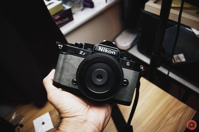 A hand holding a black Nikon Zf camera indoors near a wooden desk.