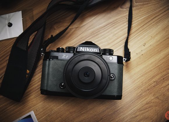 A Nikon Z fc camera with a wide lens sits on a wooden surface, strap and photo prints nearby.